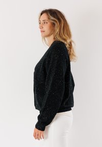 Cardigan nero lavorato a maglia con paillettes argento, vestibilità leggermente corta, maniche lunghe e polsini e bordo a costine. Indossato sopra pantaloni bianchi.