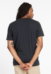 Camiseta negra de manga corta con cuello redondo, textura lisa y corte clásico, llevada por una persona de espaldas, con las manos entrelazadas detrás.