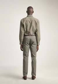 Hombre de pie de espaldas, vistiendo una camisa y pantalones verde oliva, cinturón marrón y zapatos marrones, frente a un fondo claro y sencillo.