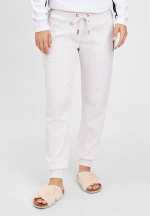 Lysegrå joggerbukser med snoretræk i taljen og ribkanter ved anklerne, båret med beige, bløde åbne tøfler på en person, der står.