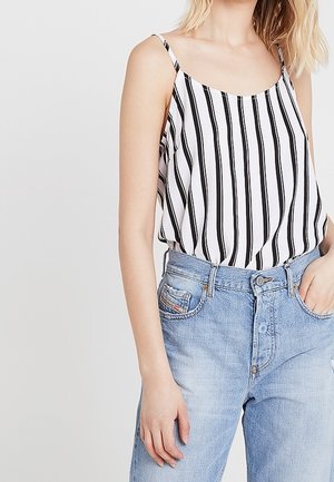 Kobieta ubrana w biały bezrękawnik z czarnymi pionowymi paskami i jasnoniebieskie dżinsy z wysokim stanem, z jedną ręką w kieszeni.