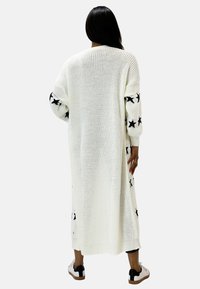 Cardigan lungo bianco lavorato a maglia con motivi di stelle nere sulle maniche e sui lati. Vestibilità ampia con orli a coste. Adatto per essere indossato a strati.