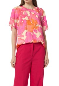 Przezroczysta bluzka z krótkim rękawem w różowo-pomarańczowy wzór kwiatowy, z marszczonym dekoltem. Połączona z jasnoróżowymi spodniami typu wide-leg.
