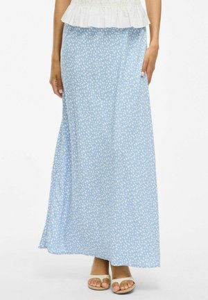 Femme portant une jupe maxi bleu clair à petits pois blancs, associée à des sandales blanches à bout ouvert et un haut blanc à volants.