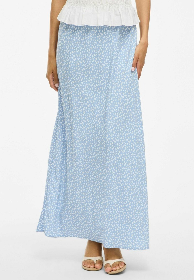 Femme portant une jupe maxi bleu clair à petits pois blancs, associée à des sandales blanches à bout ouvert et un haut blanc à volants.
