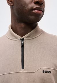 Κοντινό πλάνο ενός ατόμου που φοράει μπεζ υφασμάτινο πουκάμισο με φερμουάρ σε μαύρο χρώμα και τη λέξη "BOSS" κεντημένη στο στήθος.