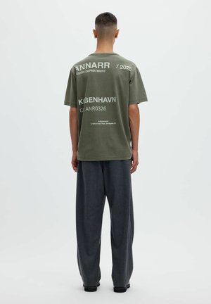 Hombre de pie de espaldas, vestido con una camiseta verde oliva con texto blanco y pantalones sueltos gris oscuro frente a un fondo liso.