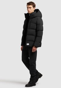 Chaqueta de plumas negra con capucha, con un diseño acolchado y cremallera frontal. Llevada sobre una camiseta negra con jeans oscuros y botas negras.