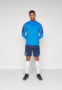 Blå atletisk top med marineblå ærmer, marineblå shorts med teal detaljer, hvide sokker og sorte fodboldsko; prydet med Nike og Chelsea logoer.