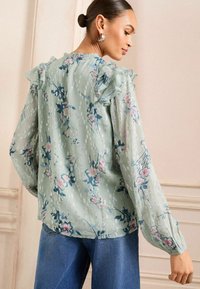 Šviesiai žalia gėlėmis puošta bluzonė su raukšlėmis ant pečių, kurią puošia rožinės ir mėlynos spalvos gėlių raštas, ilgos rankovės ir tekstūruotas paviršius.