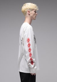 Λευκό μπλουζάκι με μακριά μανίκια που διαθέτει κόκκινα γραφικά στη αριστερή μανσέτα και μαύρο σχέδιο στο μπροστινό μέρος. Λείο βαμβακερό ύφασμα.