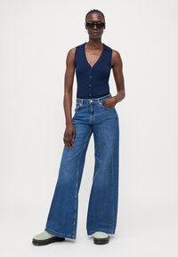 Navy ribbattu sleeveless bodysuit dengan tombol, dipadukan dengan celana jeans denim biru model wide-leg. Model mengenakan kacamata hitam dan alas kaki dengan aksen hijau.