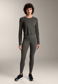 Nainen, jolla on lyhyet kiharat hiukset, pukeutuneena pitkähihamaiseen tummanharmaaseen puseroon, yhteensopiviin legginsseihin ja mustiin urheilukenkiin tasaisella harmaalla taustalla.