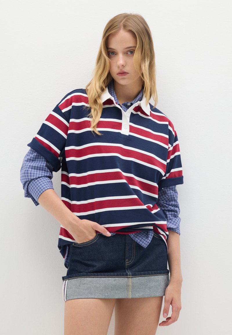 Polo de color azul marino con rayas blancas y rojas, presenta un cuello blanco. Se lleva sobre una camisa de cuadros azules y se combina con una falda corta de mezclilla.