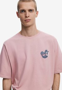 Light pink cotton t-shirt with a relaxed fit. Features a dark graphic in the shape of a heart on the left chest, textured detailing present.