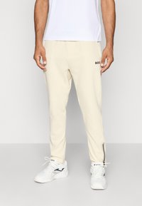 Pantalons de sport beiges avec une coupe ajustée, arborant un logo "BOSS" noir et des accents rayés près de la cheville, associés à des chaussures de sport blanches.