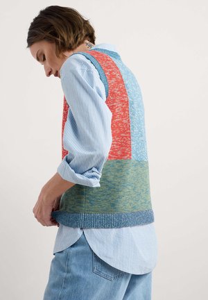 Gilet en tricot multicolore avec des blocs bleus, rouges et verts ; fil texturé ; ourlet côtelé ; superposé sur une chemise rayée bleue.