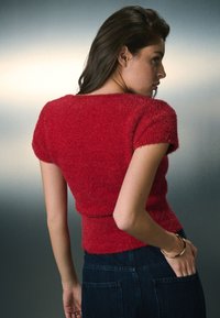 Crveni mekani pleteni džemper s kratkim rukavima i uskim krojem, uparен s tamnim trapericama. Mekana tekstura i minimalistički oblik.