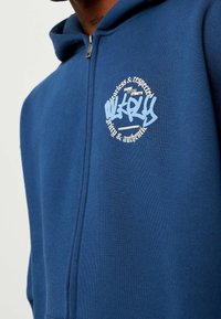 Un hoodie bleu en tissu doux, comportant une fermeture éclair à l'avant, une poche kangourou et un logo graphique blanc avec un design circulaire sur la poitrine.