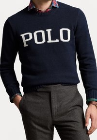 Mörkblå stickad tröja med "POLO" i vita bokstäver, rundad halsringning och ribbade muddar. Buren över en rutig skjorta i gröna, röda och blå toner.