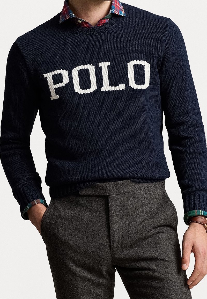 Mörkblå stickad tröja med "POLO" i vita bokstäver, rundad halsringning och ribbade muddar. Buren över en rutig skjorta i gröna, röda och blå toner.