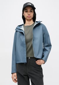 Αδιάβροχο μπουφάν ανοιχτού μπλε χρώματος με κουκούλα, με λεία υφή και φερμουάρ, συνδυασμένο με σκουρό πράσινο πουκάμισο και σκούρο παντελόνι.