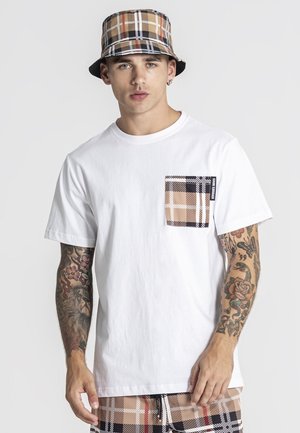 Joven con los brazos tatuados que lleva una camiseta blanca con un bolsillo a cuadros beige y negro, pantalones cortos a juego a cuadros y un sombrero tipo bucket a cuadros.
