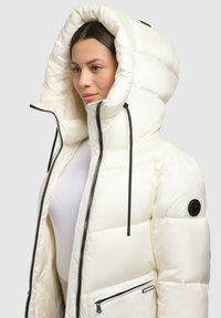 Hvid dunjakke med stor hætte, quiltet tekstur, sort lynlås og detaljer, der har en sidelomme og snoretræk ved halsen.