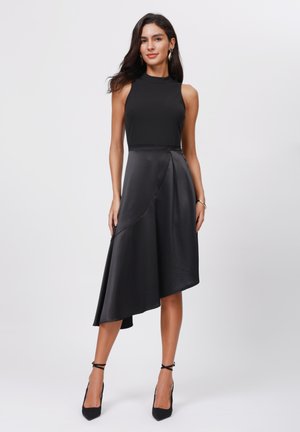 Schwarzes ärmelloses Kleid mit tailliertem Oberteil und asymmetrischem, fließendem Saum. Hergestellt aus glattem Stoff mit leichtem Glanz. Ergänzt durch schwarze High Heels.