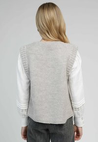 Gilet en maille grise avec un tissu texturé, accents côtelés sur les épaules, dos ouvert, porté par-dessus une blouse blanche à manches longues et un jean foncé.