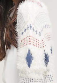 Chlupatý bílý svetr s pletenými vzory v modré a červené barvě, s texturovanými rukávy a dekorativním stehem.