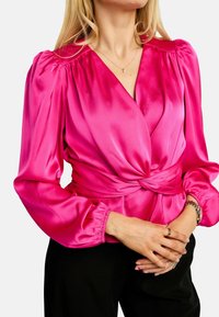 Bluza iz žameta fuksija z napihnjenimi rokavi in zavezanim pasom. Sijajna tekstura in globok V-izrez poudarjata njeno silhueto.
