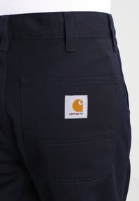 Donkerblauwe werkbroek van stevige stof, met een achterzak voorzien van een rechthoekig wit label en een gele logo. Eenvoudig ontwerp.
