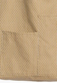 Smeđa tkanina s teksturiranim uzorkom okomitih bijelih linija. Sadrži dva džepa, jedan s lijeve strane i suptilan detalj šava.