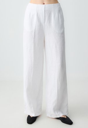 Weiße Leinen-Hose mit weitem Bein und glatter Textur, ausgestattet mit zwei vorderen Taschen und einer taillierten Taille. Kombiniert mit schwarzen flachen Schuhen.