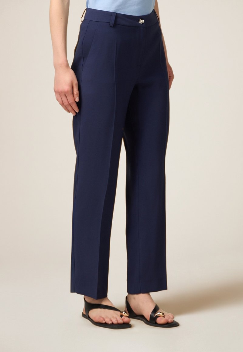Pantalon bleu marine en tissu lisse, à coupe droite, avec des plis à l'avant et une fermeture à bouton métallique.