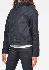 Mörk denimjacka med hög krage, framtida dragkedja och sidficka. Har en strukturerad yta och figurnära design.