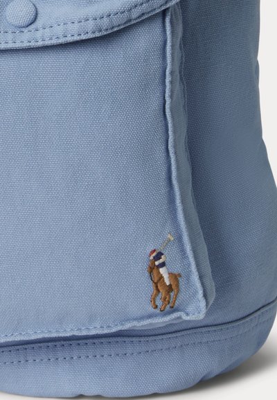 Esquina de una bolsa de tela azul claro con solapa cosida y pequeño logo bordado de un jugador de polo a caballo en varios colores.