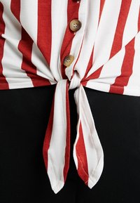 Červená a bílá pruhovaná látková košile v pase, s uzlem vpředu a třemi béžovými knoflíky. Hladká struktura proti černé látce.