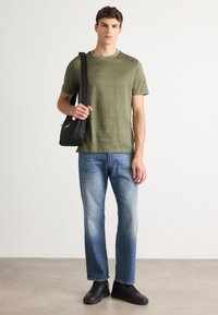 Mlad moški v olivno zeleni strukturirani majici, zbledelih modrih kavbojkah, črnih supergah in z črno torbo na rami na svetli podlagi.