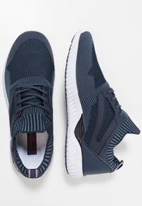 Sneakers in maglina blu navy con suole bianche, polsini a righe, design con lacci e motivi testurizzati mostrati da vedute superiori e laterali.