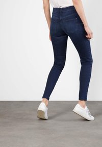 Persona vestida con jeans ajustados azul oscuro y zapatillas blancas, de pie sobre un suelo gris contra un fondo blanco, vista desde atrás.