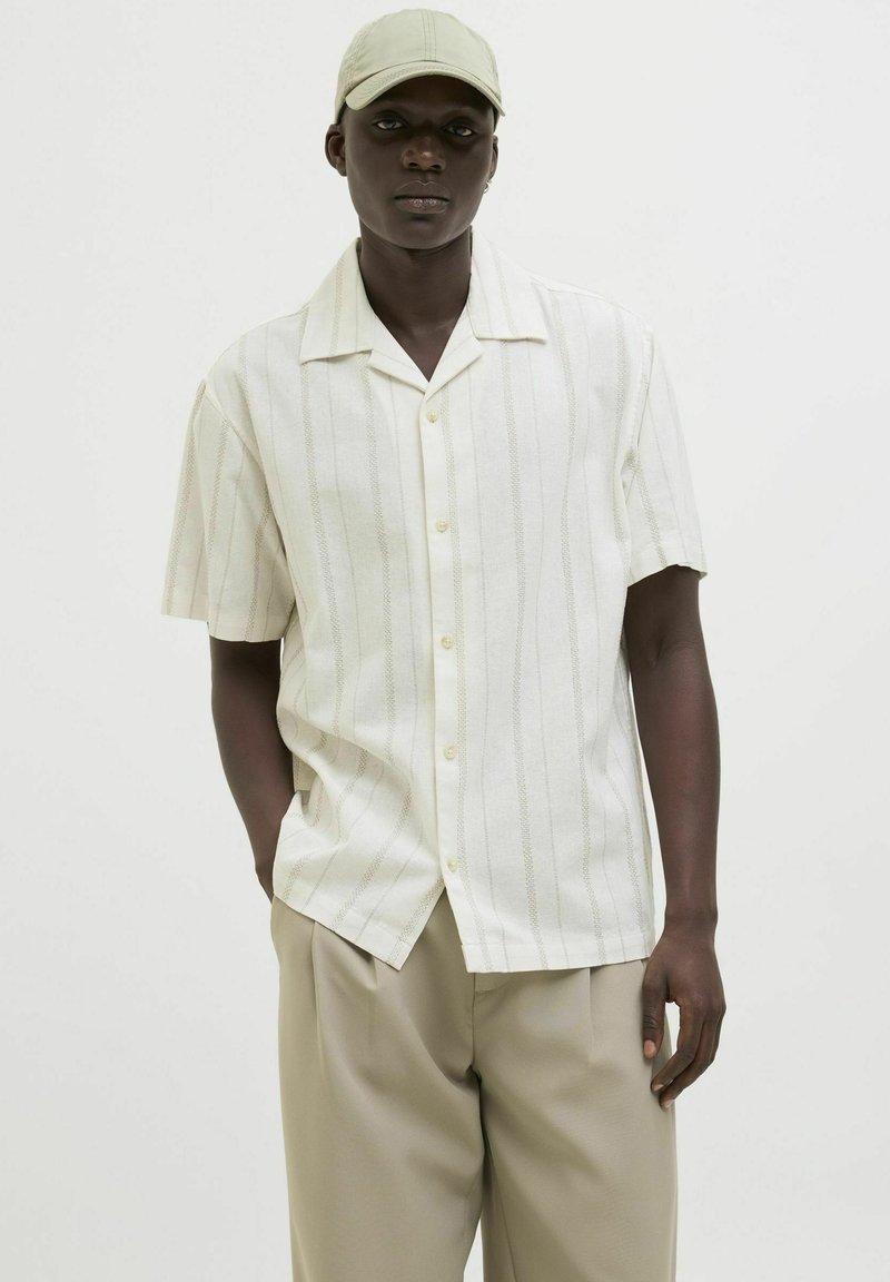 Chemise à manches courtes, à col, de couleur crème avec des rayures verticales beige clair. Comprend des boutons et une coupe décontractée. Associée à un pantalon beige.