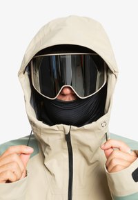 Giacca impermeabile beige con cappuccio, dotata di un colletto alto, gaiter per il collo nero e occhiali protettivi a specchio oversize. Dettaglio verde sulle maniche.