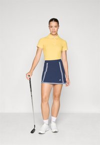 Polo amarillo de canalé con cuello, combinado con una falda corta azul marino con rayas blancas y un logo. Sosteniendo un palo de golf; zapatillas y calcetines blancos.