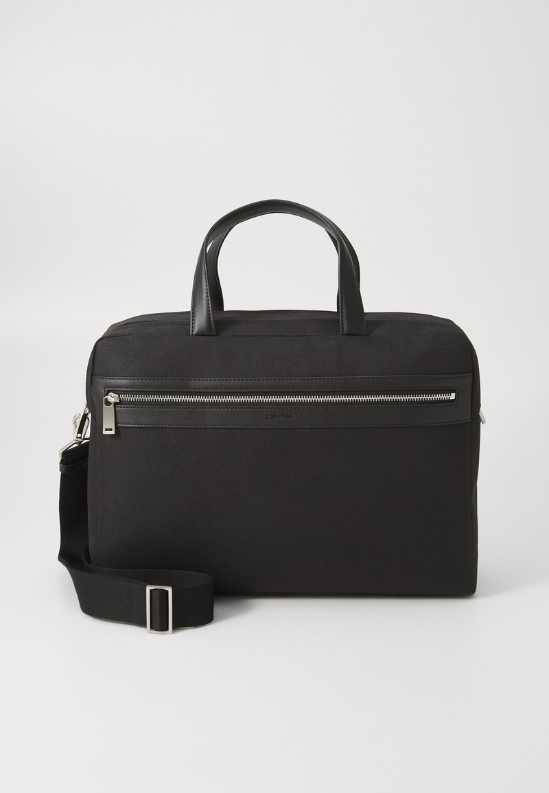 Bolsa de tecido preta com acabamentos em pele, apresentando uma pega superior, correia de ombro removível, bolso frontal com fecho e forma retangular.