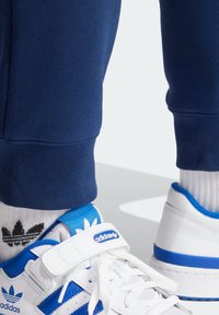 White sneakers with blue accents, featuring a textured upper, adjustable strap, and three stripes design. Worn with navy joggers and white socks.