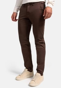 Chinos slim-fit marrones con una textura sutil, que cuentan con cremallera, bolsillos y pasadores para cinturón, combinados con zapatillas beige.