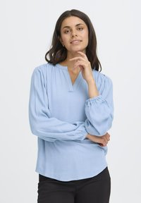 Femme aux cheveux bruns portant un blouse à manches longues bleu clair avec des poignets froncés, posant avec une main près de son visage sur un fond uni.