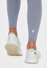 Białe buty sportowe o fakturowanej powierzchni, szare legginsy o dopasowanym kroju, z logo w okolicy kostki.